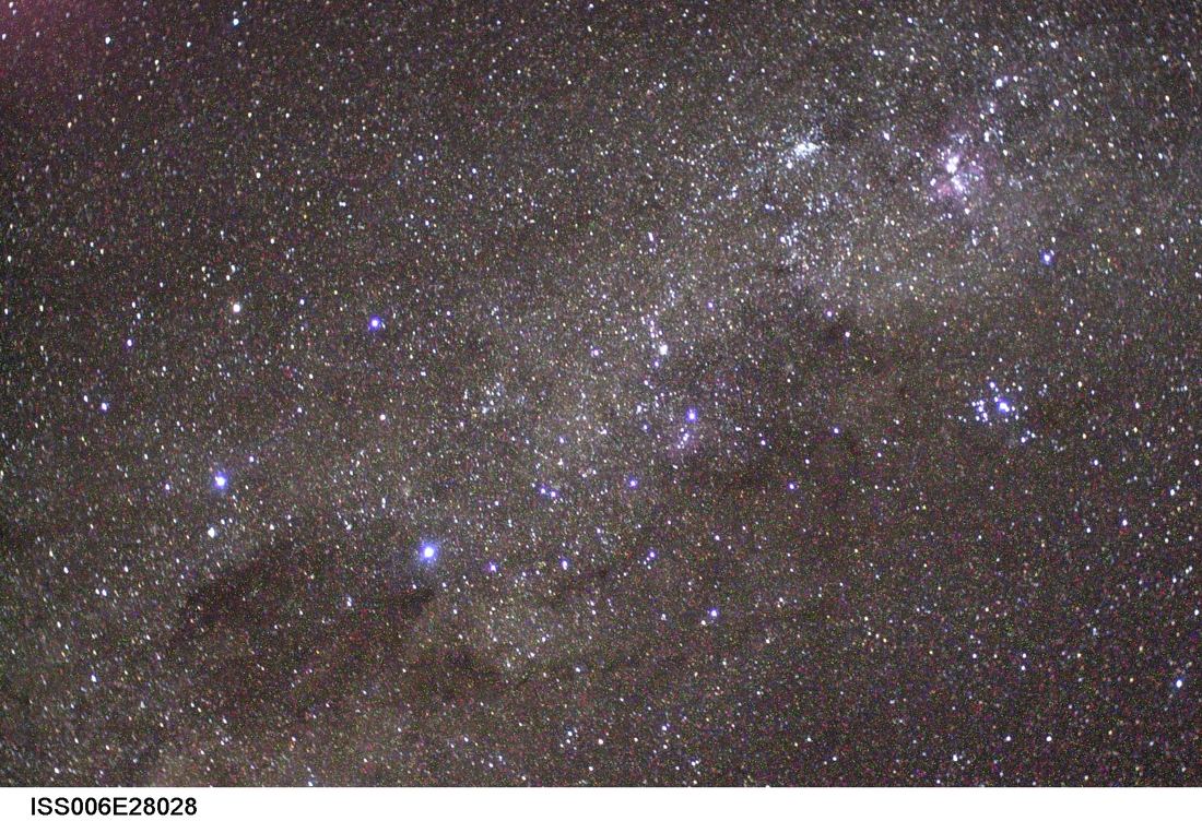 Fotografia de campo estelar feita do espaço: faixa brilhante da Via Láctea com a pequena cruz de quatro estrelas do Cruzeiro do Sul e uma mancha escura ao lado.