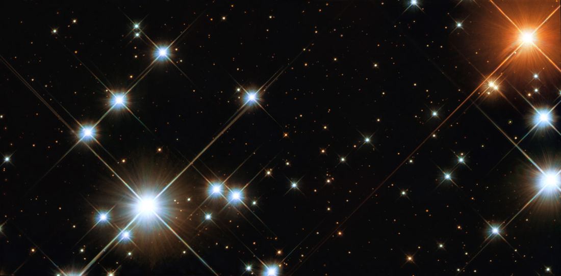 Várias estrelas azuis muito brilhantes com picos de difração, uma estrela vermelho-alaranjada intensa e muitas estrelas fracas coloridas sobre fundo escuro.