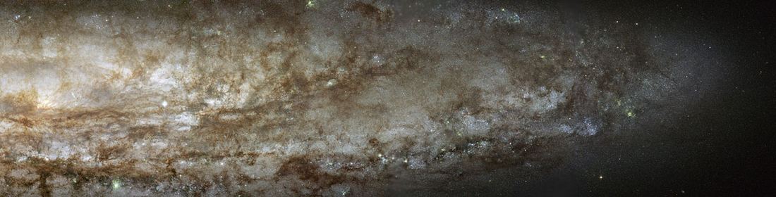 Galáxia espiral fortemente inclinada, com disco alongado cheio de pontos azuis de estrelas jovens, nós rosados de gás e filamentos escuros de poeira sobre fundo estrelado.