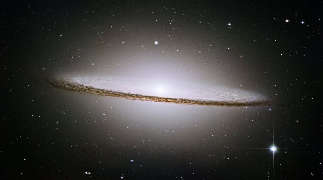 Galáxia vista de lado, com um bojo central branco e luminoso e um anel fino e escuro de poeira que o atravessa horizontalmente, sobre fundo preto com estrelas.