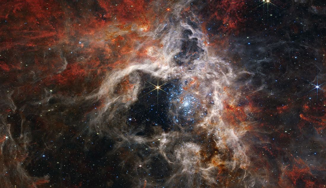 Nuvem de gás em tons de azul, laranja e marrom com filamentos que lembram pernas de aranha e um aglomerado denso de estrelas azuis brilhantes no centro.