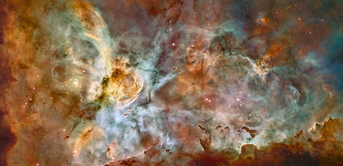 Panorama de nuvens de gás em tons de laranja, marrom e azul, com pilares escuros de poeira e muitas estrelas brilhantes espalhadas.