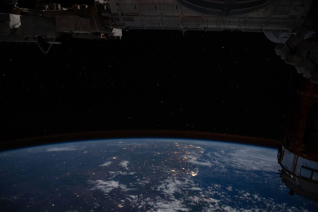 Vista noturna da Terra a partir da órbita, com luzes de cidades no litoral brasileiro, o horizonte curvo e estrelas no céu escuro acima.