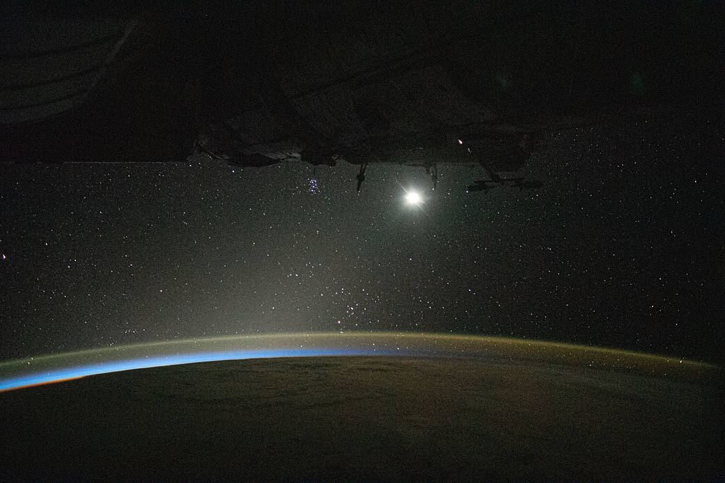 Horizonte curvo da Terra com brilho azul da atmosfera, luz da Lua à esquerda e faixa da Via Láctea no céu escuro acima.