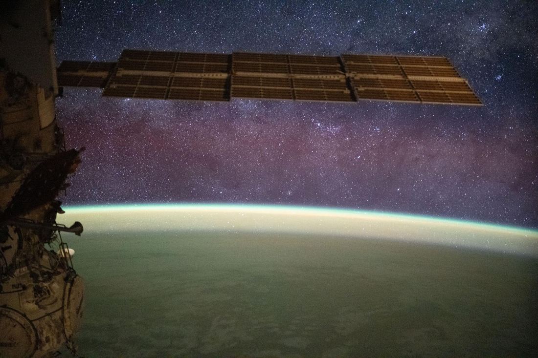 Fotografia noturna feita do espaço mostrando a faixa esbranquiçada da Via Láctea cruzando o céu acima da curva da Terra, com um brilho amarelo-esverdeado na borda do planeta.